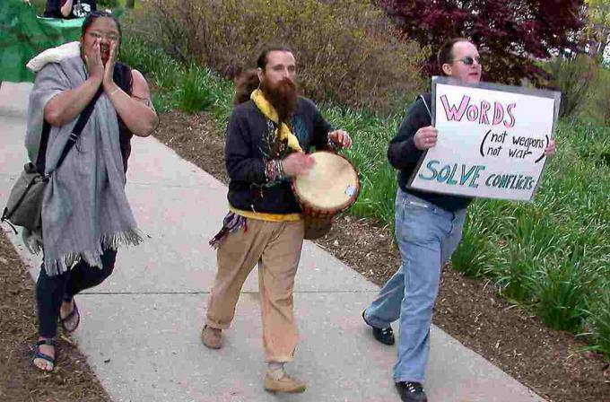 Una donna urla, un uomo barbuto suona un tamburo e un altro uomo tiene un cartello di protesta.