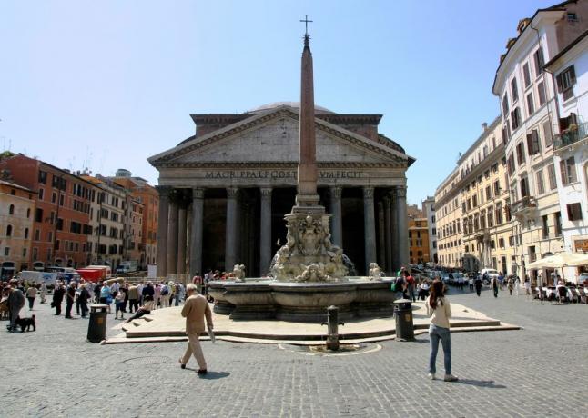 turisti che camminano sulla piazza di pietra vicino alla fontana con piccola torre e croce cristiana con circostanti edifici in pietra