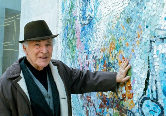 L'artista Marc Chagall che indossa un cappello preme la mano contro un muro con disegni a mosaico blu.